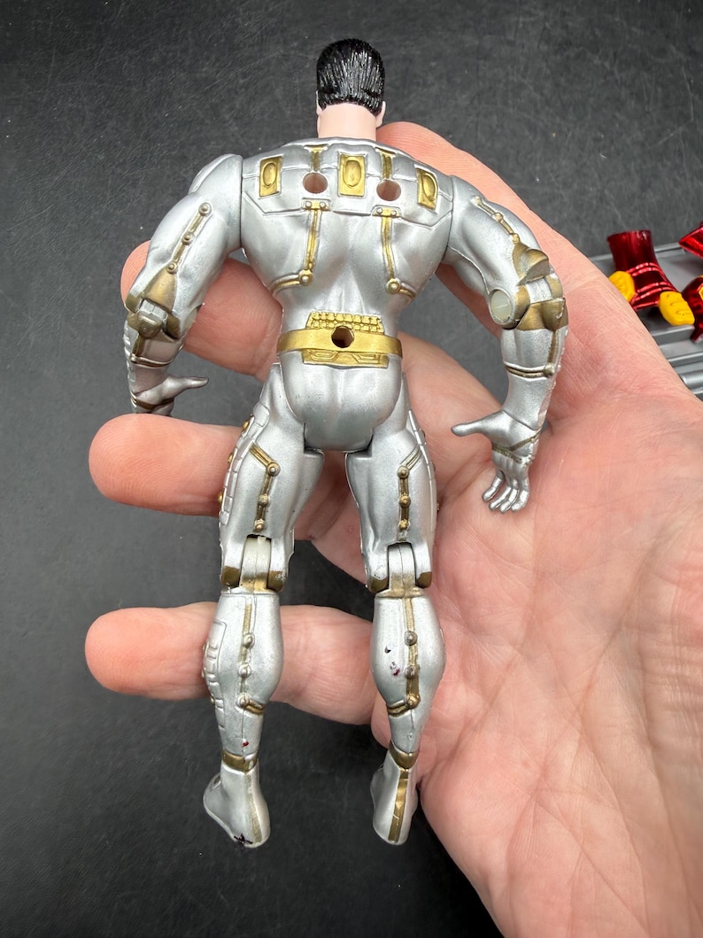 May include: Silver action figure with gold accents, viewed from the back. The figure has a black head and is articulated at the joints. The figure is held in a hand.