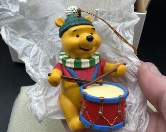 Puede incluir: Adorno amarillo de Winnie the Pooh con gorro verde y atuendo rojo y azul, tocando un tambor rojo con baquetas marrones. El adorno tiene una cuerda dorada para colgar.