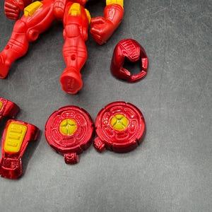 May include: A disassembled Iron Man action figure in red and yellow. The figure's torso, legs, arms, and head are separated. Additional components include two circular chest pieces and a helmet. The figure is on a gray surface.