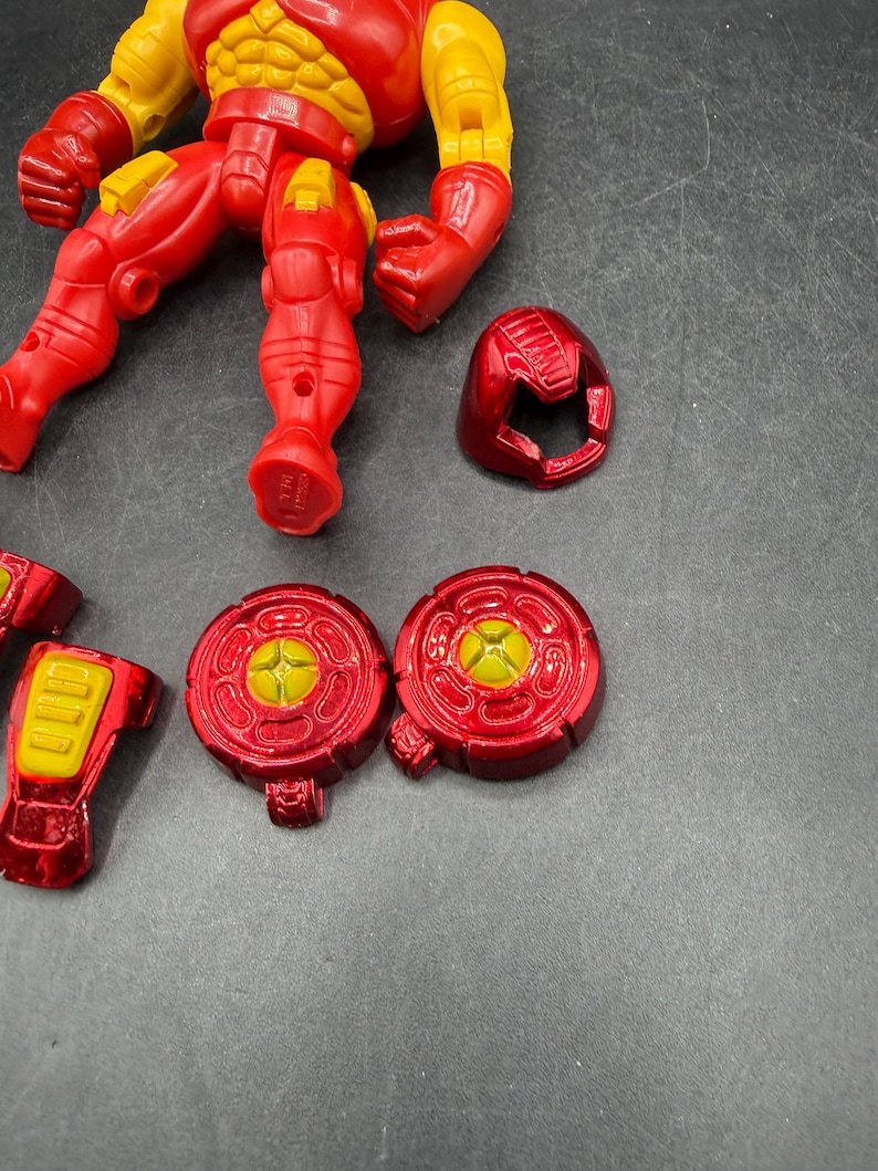 May include: A disassembled Iron Man action figure in red and yellow. The figure's torso, legs, arms, and head are separated. Additional components include two circular chest pieces and a helmet. The figure is on a gray surface.