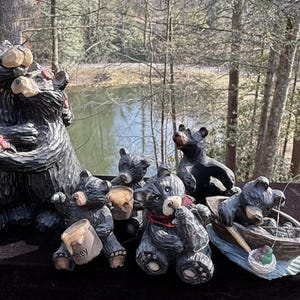 May include: A collection of black bear figurines, some holding honey pots, others in a boat fishing. One bear hugs another, holding flowers. The figurines are carved with a rustic, textured finish, set against a backdrop of trees and water.