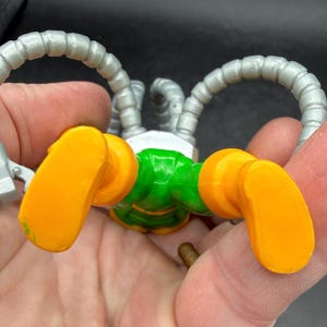 Puede incluir: Un robot de juguete con cuerpo verde, pies naranjas y brazos plateados. Los brazos del robot son curvos y terminan en forma de garra. Los pies son de color naranja brillante.