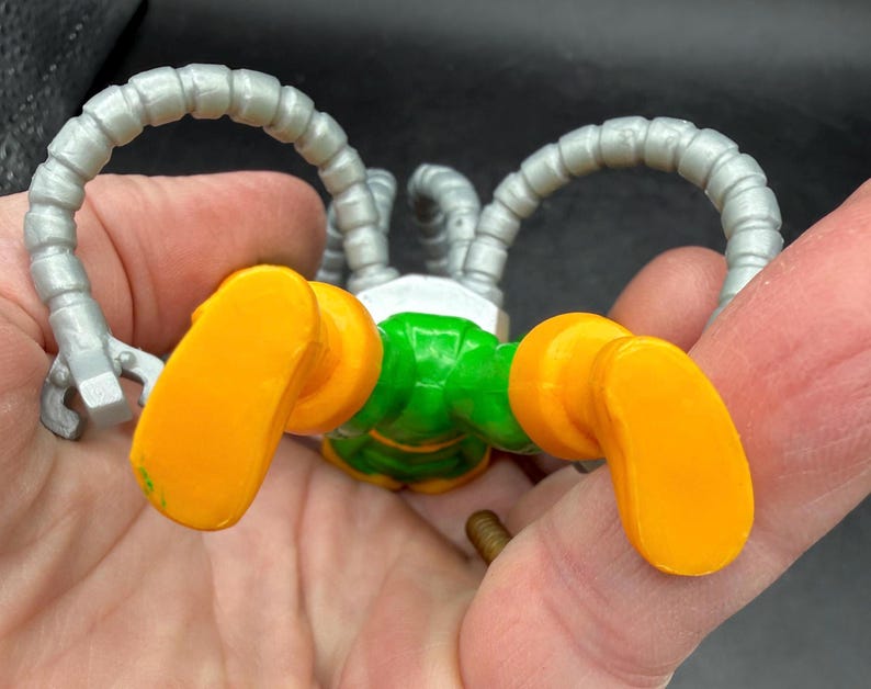 Puede incluir: Un robot de juguete con cuerpo verde, pies naranjas y brazos plateados. Los brazos del robot son curvos y terminan en forma de garra. Los pies son de color naranja brillante.