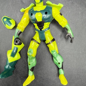 May include: A yellow and green action figure with a futuristic design. The figure has a helmet, shoulder pads, and articulated limbs. The figure is missing a piece from its left arm. The figure is made of plastic.
