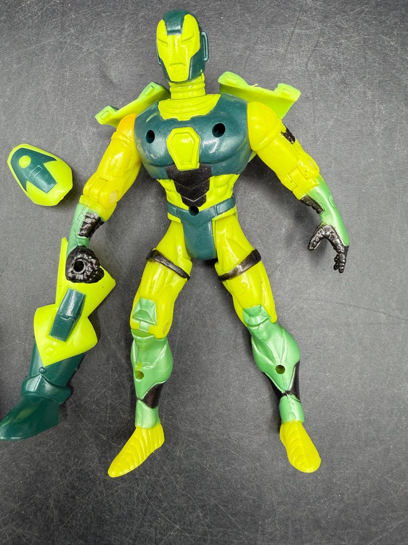 May include: A yellow and green action figure with a futuristic design. The figure has a helmet, shoulder pads, and articulated limbs. The figure is missing a piece from its left arm. The figure is made of plastic.