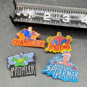 Może przedstawiać: Cztery kolorowe przypinki z postaciami superbohater&oacute;w, w tym Chameleon, Spider-Man, Cyborg Spider-Man i The Prowler, są wyświetlane w pobliżu taśmy mierniczej. Taśma pokazuje pomiary w centymetrach.