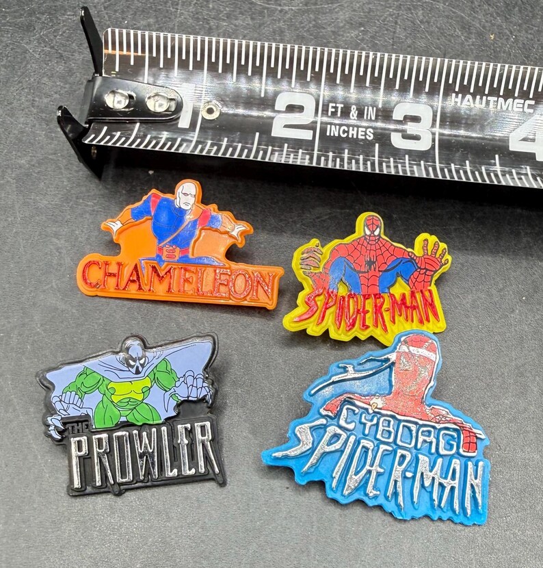 Może przedstawiać: Cztery kolorowe przypinki z postaciami superbohater&oacute;w, w tym Chameleon, Spider-Man, Cyborg Spider-Man i The Prowler, są wyświetlane w pobliżu taśmy mierniczej. Taśma pokazuje pomiary w centymetrach.