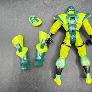 May include: A yellow and green action figure with detachable parts. The figure has a helmet, shoulder pads, and leg armor. The figure's hands are black. The figure is on a dark surface.