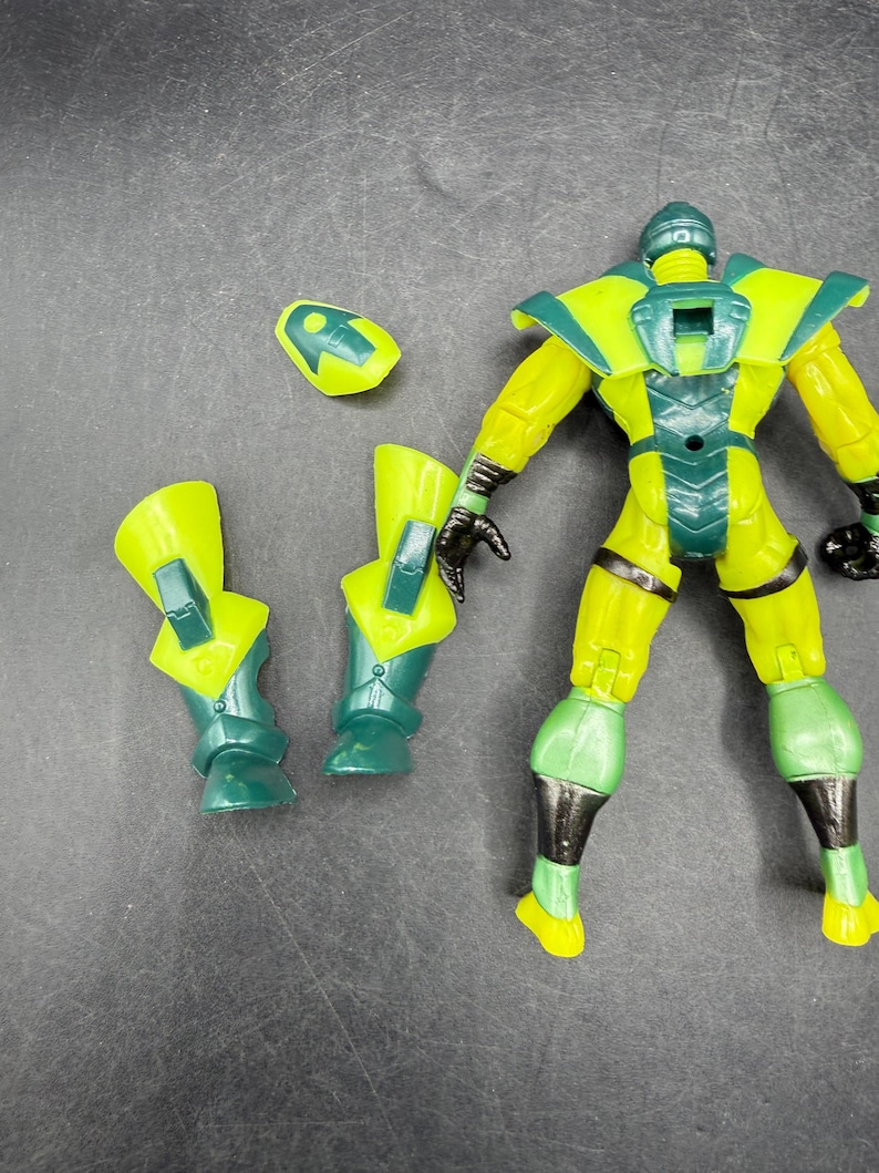 May include: A yellow and green action figure with detachable parts. The figure has a helmet, shoulder pads, and leg armor. The figure's hands are black. The figure is on a dark surface.