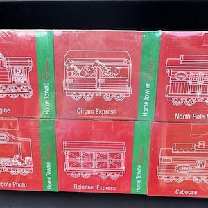 May include: A set of eight red and green boxes with white outlines of train cars. Each box features a different train car design, including an engine, a circus express, a reindeer express, a caboose, and a north pole mail car. The boxes are labeled with the names of the train cars and the words "Home Towne".