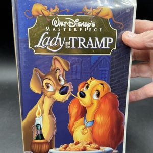 May include: A VHS tape of Disney's animated film "Lady and the Tramp." The cover shows Lady and Tramp sharing spaghetti, with a candle and a bottle of wine on the table. The title is in gold lettering, with the words "Walt Disney's Masterpiece" above.