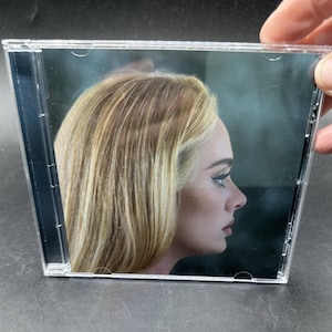 May include: A clear plastic CD jewel case featuring a profile portrait of a person with long blonde hair and dark eyeliner against a dark background. The cover art shows the side of the face looking toward the right edge of the square case.