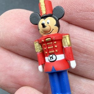 以下が含まれることがあります： おもちゃの兵隊の衣装を着た小さなミッキーマウスのフィギュア。赤いジャケットに金色のアクセント、青いズボン、黒いブーツ、金色のストライプが入った赤い帽子を着用。文字「M」が描かれた円形のバックルが付いた白いベルトが見える。