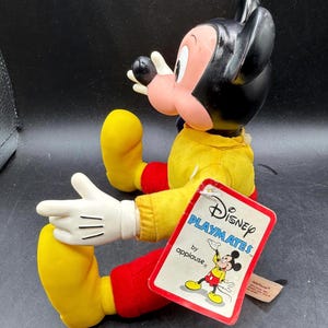 May include: A vintage Mickey Mouse doll by Applause. The doll has a black head, pink face, and wears a yellow shirt, red pants, and yellow shoes. The doll's tag reads "Disney Playmates by applause."