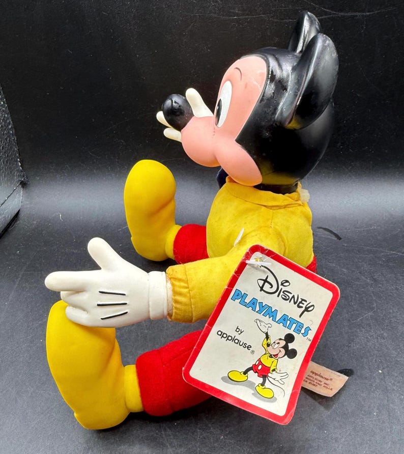 May include: A vintage Mickey Mouse doll by Applause. The doll has a black head, pink face, and wears a yellow shirt, red pants, and yellow shoes. The doll's tag reads "Disney Playmates by applause."