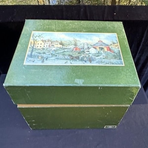 May include: A green cardboard box with a hinged lid. The lid features a colorful illustration of a rural scene with houses, a barn, and a windmill. The box is closed.