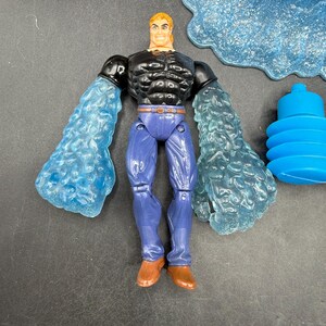 May include: Action figure of a muscular man with blonde hair, wearing a black top, blue pants, and brown boots. The figure has translucent blue water-like arms. Additional blue water-themed accessories are included.