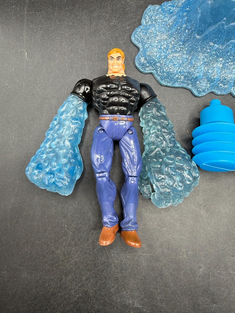 May include: Action figure of a muscular man with blonde hair, wearing a black top, blue pants, and brown boots. The figure has translucent blue water-like arms. Additional blue water-themed accessories are included.