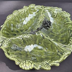 May include: A green ceramic serving dish with three sections. The dish is decorated with pine cone and leaf details around the rim and in the center. The dish has a glossy finish and is set against a dark background.