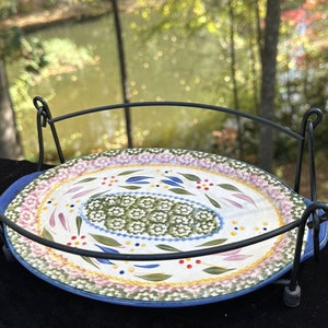 Könnte beinhalten: Eine farbenfrohe ovale Keramik-Servierplatte mit einem Blumendesign in den Farben Rosa, Grün, Blau und Gelb. Die Platte steht auf einem schwarzen Metallständer mit einem Drahtgestell.