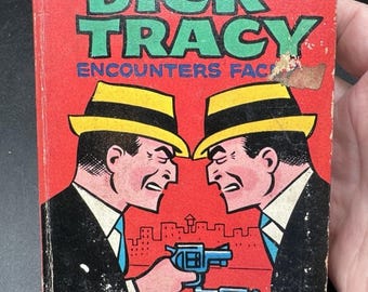 Puede incluir: Cómic vintage "Dick Tracy Encounters Face". La portada muestra a dos hombres con trajes y sombreros amarillos apuntándose revólveres azules sobre un fondo rojo. El título es verde.