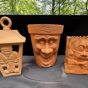 May include: Three terracotta planters with carved faces. A lantern-shaped planter features a carved figure. A large planter has a smiling face. A third planter is shaped like a paper bag with a smiling face. All are in shades of brown.