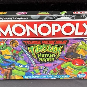 Può includere: Scatola del gioco da tavolo Monopoly con Teenage Mutant Ninja Turtles: Mutant Mayhem. La scatola presenta il logo Monopoly rosso e bianco, con i personaggi TMNT in pose d'azione su uno sfondo di muro di mattoni viola. Il testo recita "Combatti i mutanti con i tuoi poteri da tartaruga!"