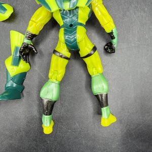 May include: A yellow and green action figure with black accents. The figure has a helmet, shoulder pads, and a chest plate. The figure's limbs are articulated, and it appears to be a superhero or villain character.