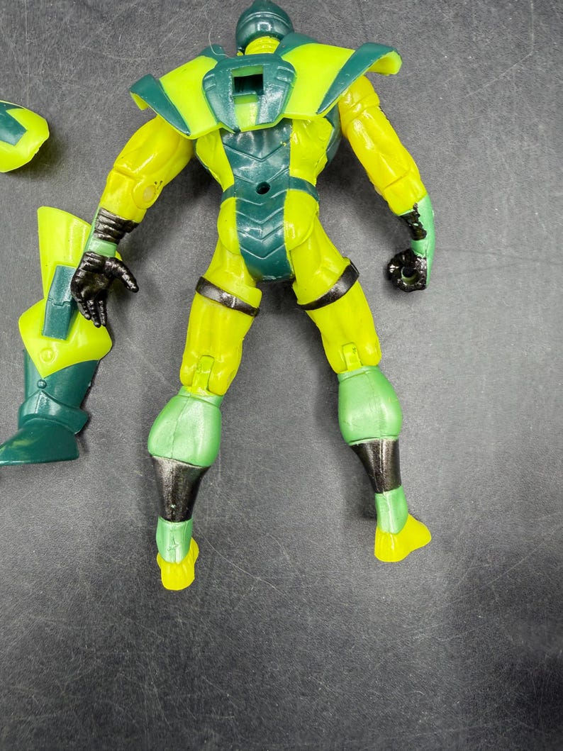 May include: A yellow and green action figure with black accents. The figure has a helmet, shoulder pads, and a chest plate. The figure's limbs are articulated, and it appears to be a superhero or villain character.