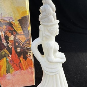 May include: White porcelain figurine of a woman wearing a large hat with a ruffled skirt. The figurine is a vintage perfume bottle.