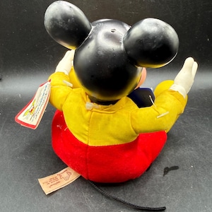 May include: A vintage Mickey Mouse plush doll, featuring black ears, a yellow jacket, red shorts, and white gloves. The doll has a black tail and a tag attached. A classic Disney collectible.