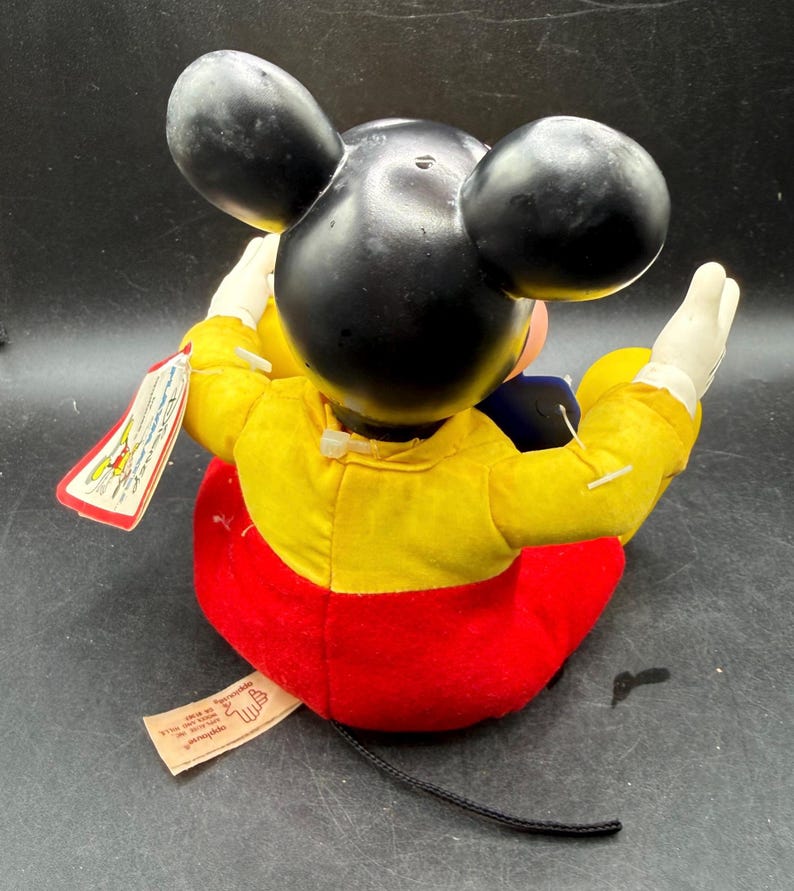 May include: A vintage Mickey Mouse plush doll, featuring black ears, a yellow jacket, red shorts, and white gloves. The doll has a black tail and a tag attached. A classic Disney collectible.