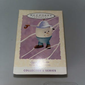 May include: A collectible "Keepsake Ornament" box featuring an egg character dressed in golf attire. The egg is wearing a blue cap with "Golf '94" on it, blue pants, and brown shoes. The box has a purple background with white lines.