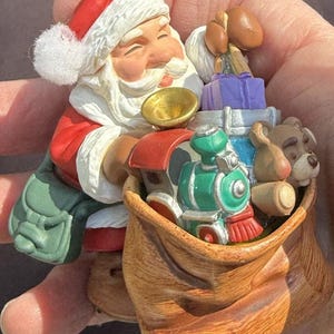 Könnte beinhalten: Ein Weihnachtsschmuck mit dem Weihnachtsmann in rotem Anzug und Hut, der ein goldenes Horn hält. Ein brauner Sack ist mit Geschenken gefüllt, darunter ein grüner Zug, ein Teddybär und verpackte Geschenke. Der Schmuck ist detailliert und festlich.