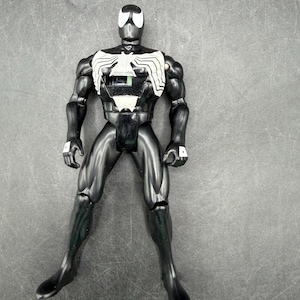 May include: Black action figure of a superhero with a white spider emblem on the chest and a white spider-shaped design on the head. The figure is articulated and stands with legs apart.