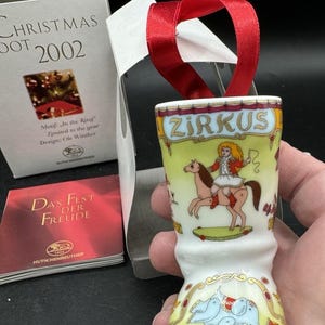 May include: A white ceramic Christmas ornament with a red ribbon. The ornament features a circus theme with the word "ZIRKUS" at the top. Illustrations include a girl riding a horse and elephants. The box reads "CHRISTMAS DOT 2002".