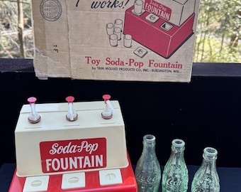 May include: Vintage toy soda pop fountain with three red and white dispensers and a red base. The fountain has a white sign that reads "Soda-Pop Fountain". There are four clear glass bottles with the Coca-Cola logo.