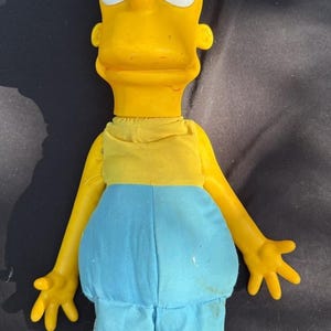 May include: A Bart Simpson doll, featuring a yellow head with spiky hair, large eyes, and a prominent nose. The doll wears a yellow shirt, blue shorts, and blue shoes. The doll is a collectible item.
