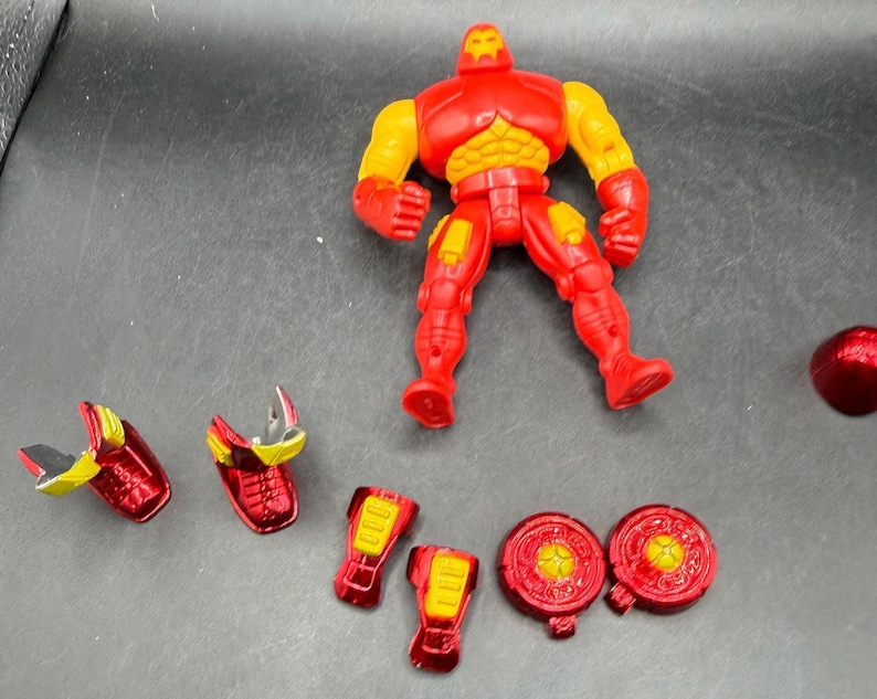 May include: A red and yellow Iron Man action figure with detachable armor pieces. The figure is standing, with various armor components scattered around, including shoulder pads, leg pieces, and chest plates. The figure's design is based on the Marvel Comics character.