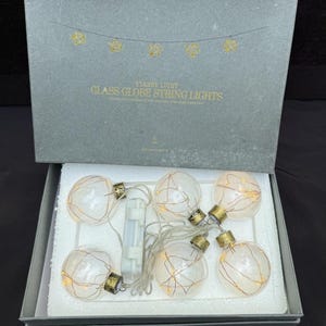 May include: A gray box containing glass globe string lights with gold-toned caps. The lid features the text STARRY LIGHT GLASS GLOBE STRING LIGHTS and a graphic of lights. Inside, six clear glass globes with internal copper wire lights are arranged.