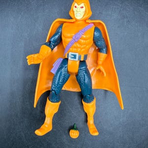 May include: Orange and blue action figure of a character with a hooded cowl and cape. The figure has orange boots, gloves, and a belt. A small pumpkin accessory is included. The figure is posed standing upright.