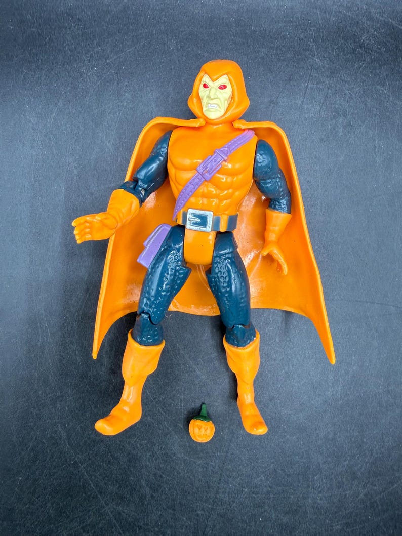 May include: Orange and blue action figure of a character with a hooded cowl and cape. The figure has orange boots, gloves, and a belt. A small pumpkin accessory is included. The figure is posed standing upright.