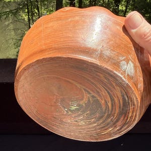 May include: A handcrafted, reddish-brown wooden bowl with a wavy rim. The bowl's exterior shows a wood grain pattern, while the interior has a swirling, glossy finish. The bowl is held by a hand, showcasing its size and shape.