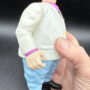 May include: A figurine of a bald man in a white suit with a purple collar and cuffs, light blue pants, and black shoes. The figure is being held by a hand.
