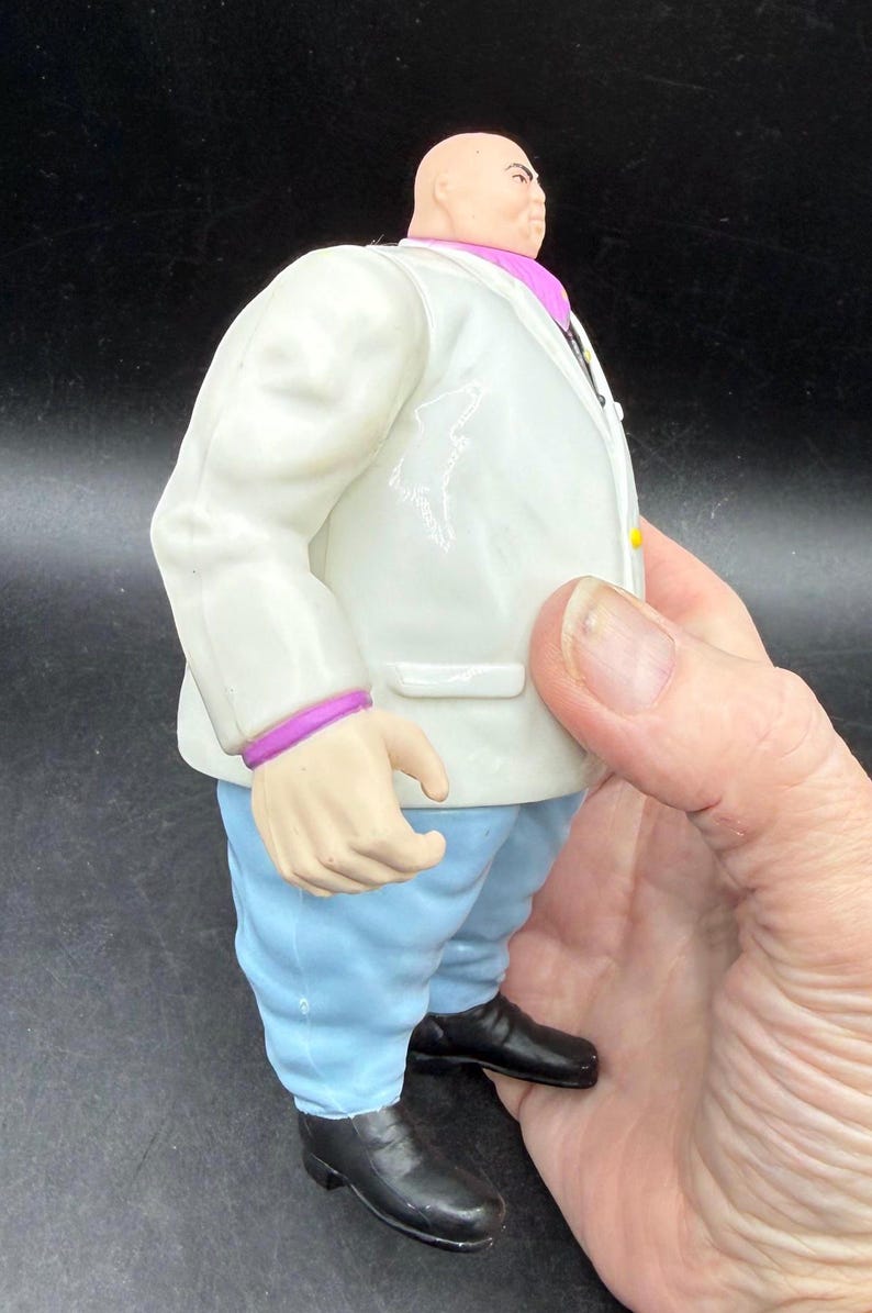 May include: A figurine of a bald man in a white suit with a purple collar and cuffs, light blue pants, and black shoes. The figure is being held by a hand.