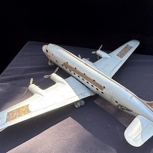 May include: A vintage toy aeroplane model made of metal, painted in a silver colour with brown accents. The model has a long fuselage, two wings, and a tail. The aeroplane has a propeller on each wing and a small window on the side of the fuselage.