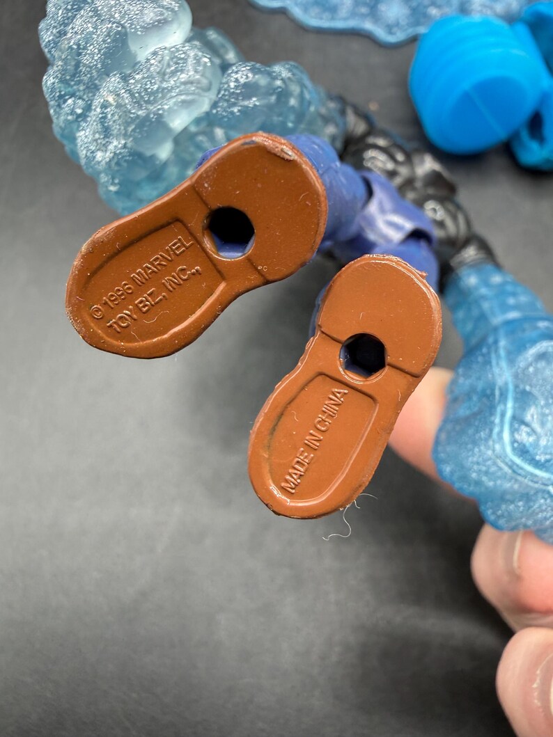 May include: Close-up of the feet of a blue action figure with brown soles. The soles are stamped with "&copy; 1996 MARVEL TOY BIZ, INC." and "MADE IN CHINA". The figure has blue and clear plastic ice-like attachments.