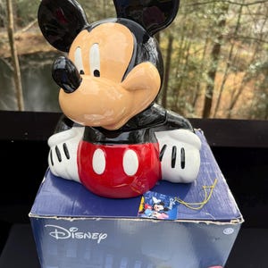 May include: A ceramic Mickey Mouse figurine with a black head and ears, a tan face, and a red and white torso. The figurine is on top of a blue Disney-branded box. The box features illustrations of Mickey Mouse and Minnie Mouse.