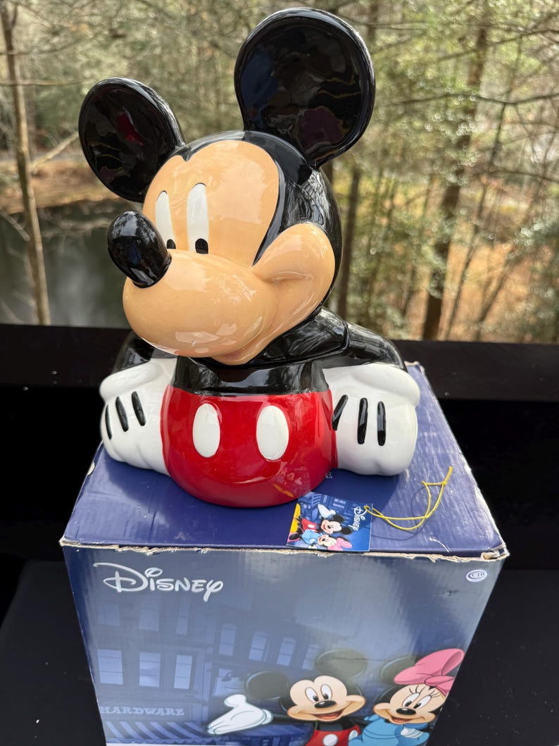May include: A ceramic Mickey Mouse figurine with a black head and ears, a tan face, and a red and white torso. The figurine is on top of a blue Disney-branded box. The box features illustrations of Mickey Mouse and Minnie Mouse.