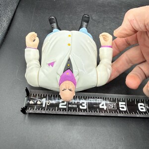 May include: A vintage action figure of a character in a white coat, purple tie, and blue pants. The figure is approximately 3 inches long. The figure has black shoes and is lying on its back.